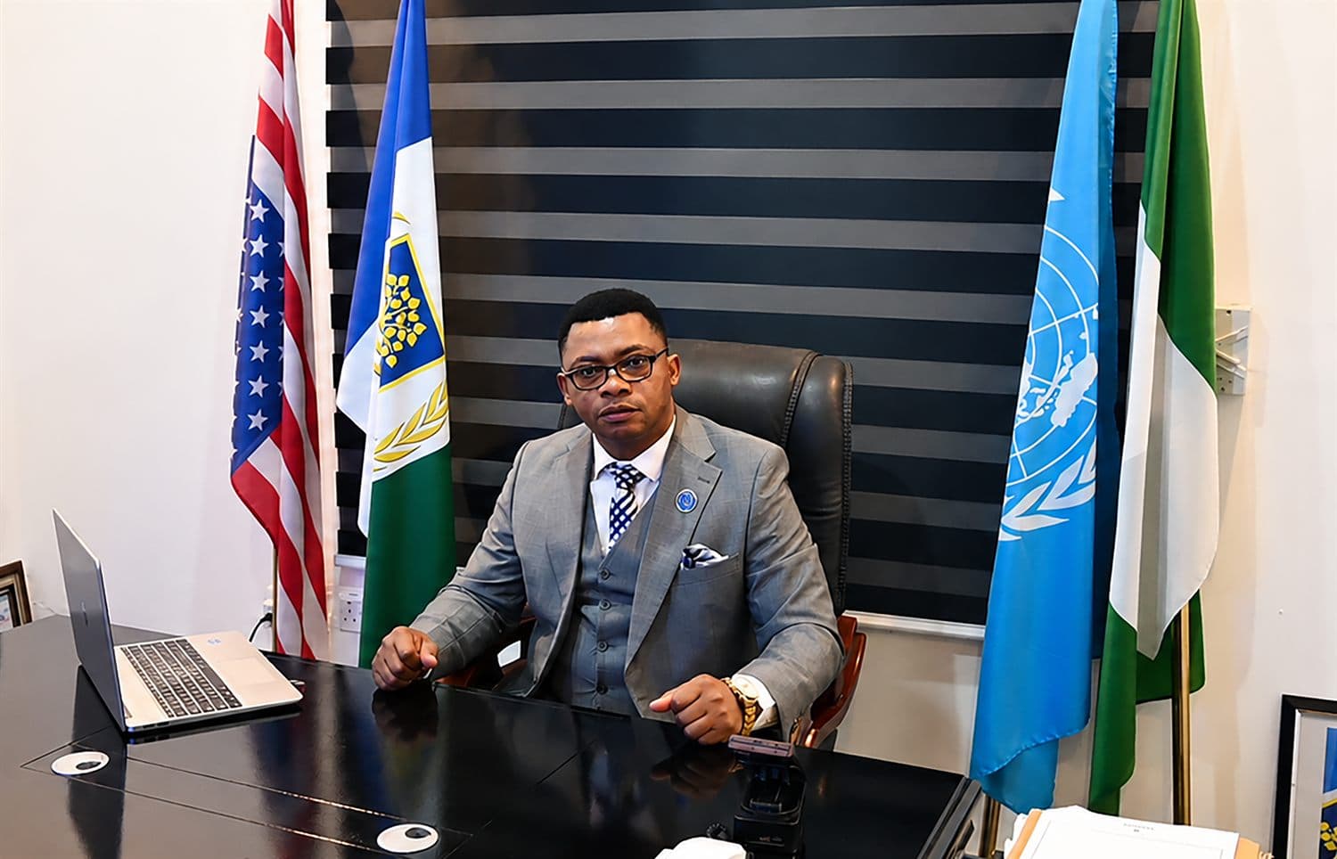 Joseph Legend Mfon at his desk, flanked by the flags of the United States, the United Nations and Nigeria.