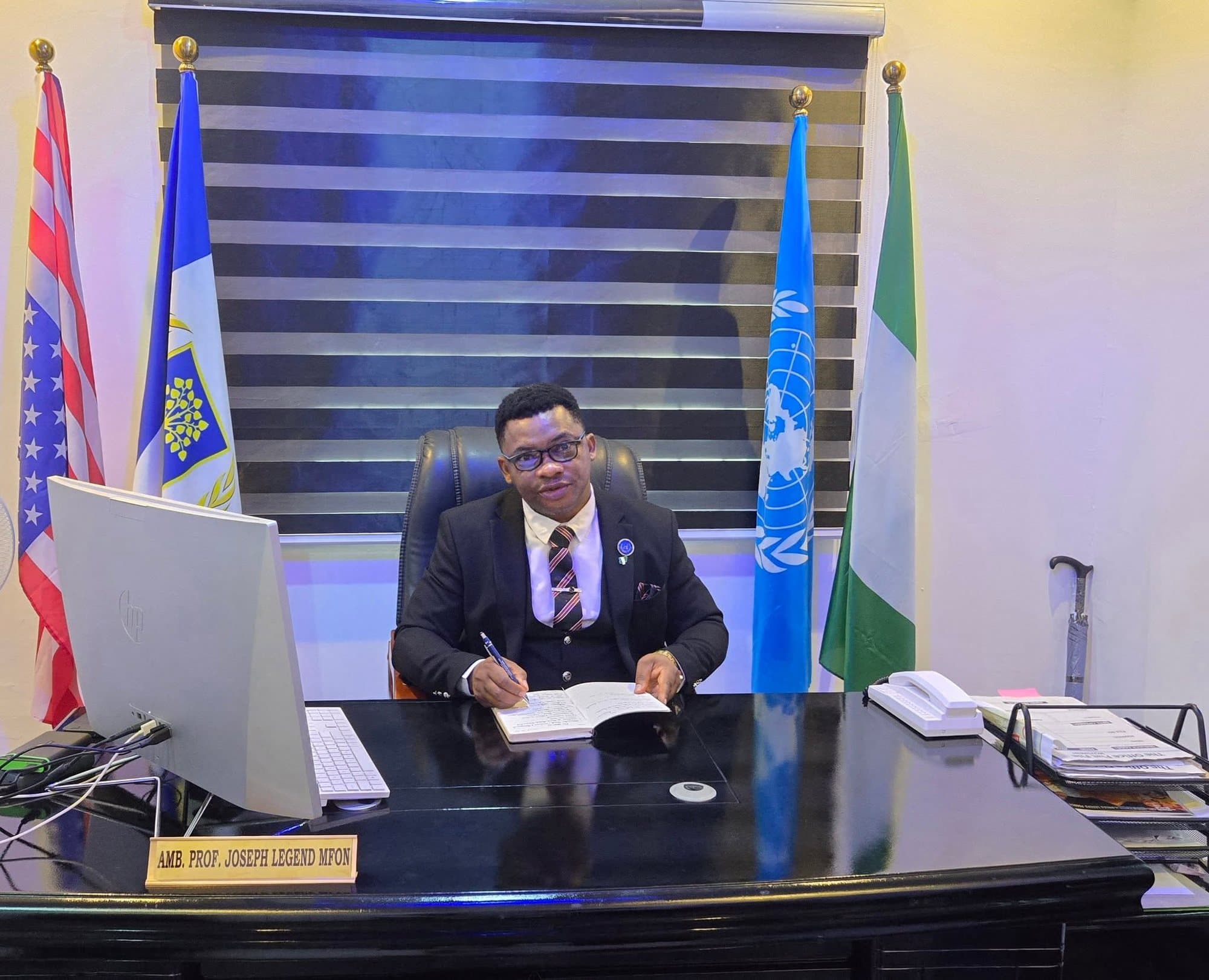 Joseph Legend Mfon at his desk, flanked by the flags of the United States, the United Nations and Nigeria.